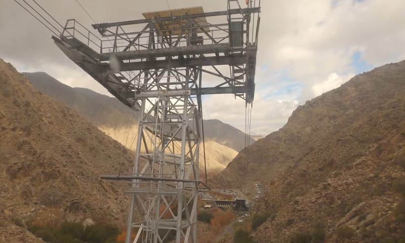 A steel cableway tower and suspended cables span a rugged mountain valley beneath a cloudy sky
