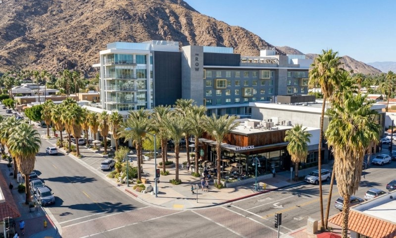 Modern glass-and-stone hotel complex lines a palm tree–shaded street beneath rugged desert mountains